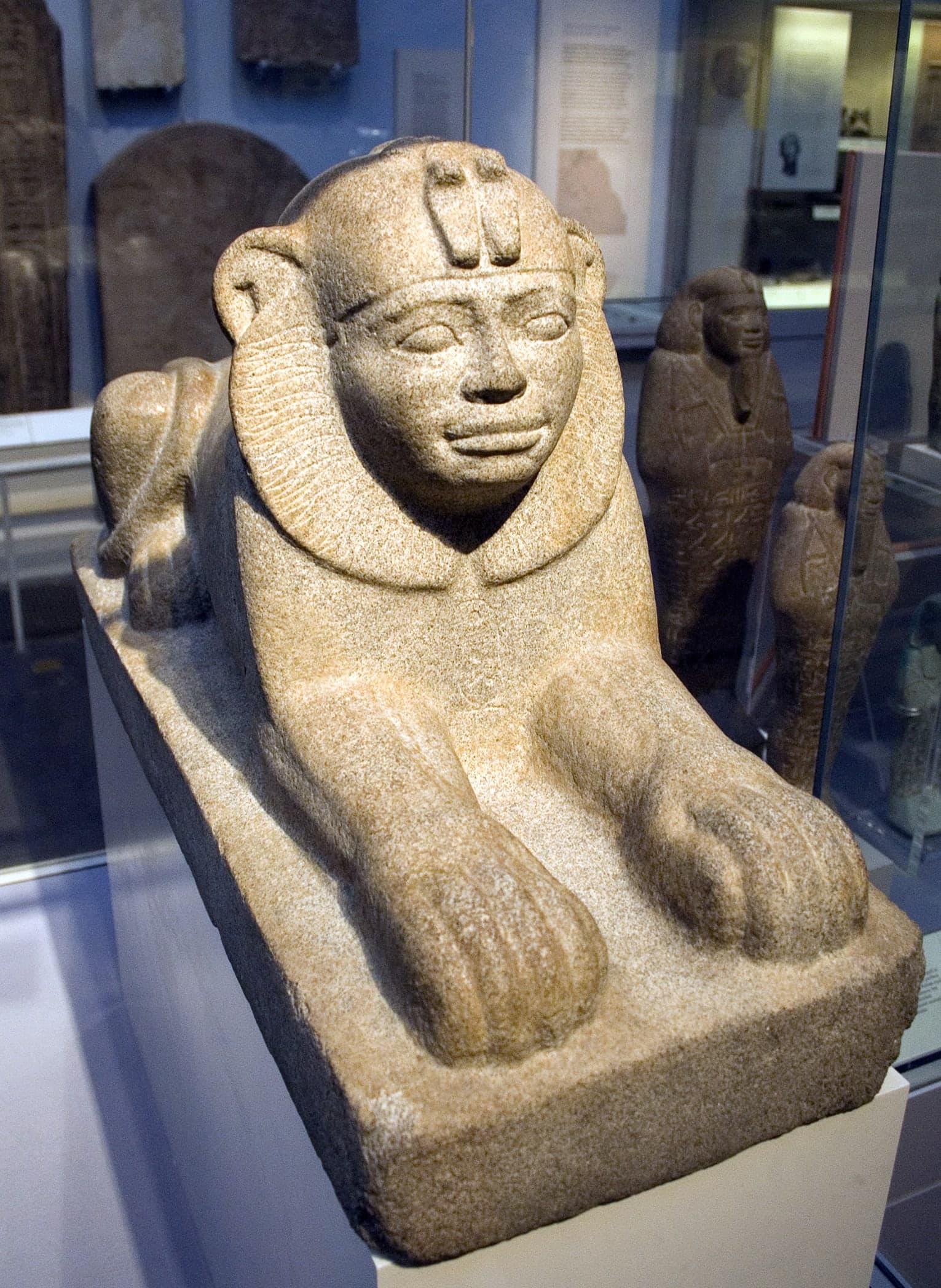 A small recumbent granite sphinx with a lion’s body and a human head bearing full Kushite royal features, the stone a sandy grey, hieroglyphs visible on the surface.