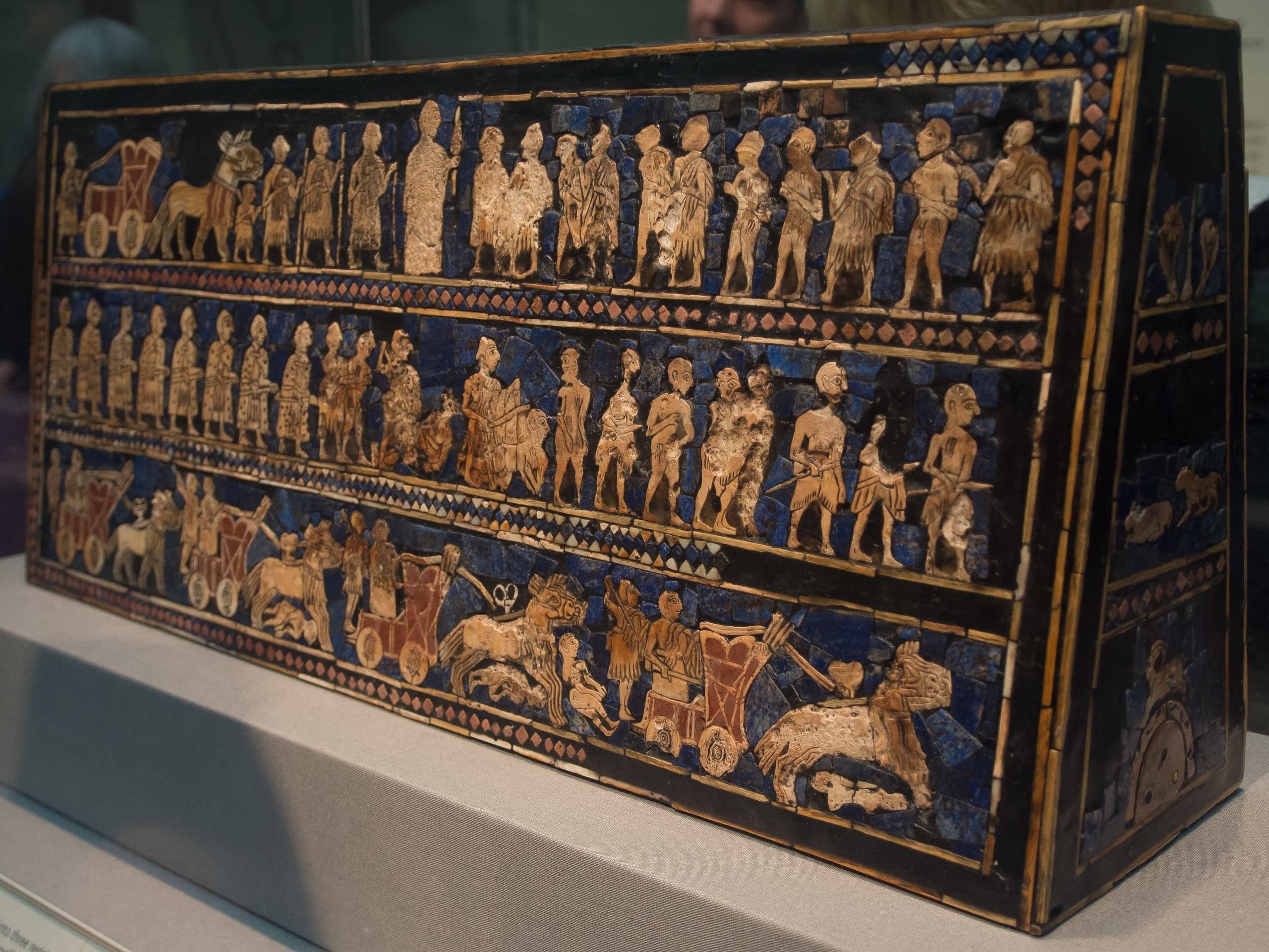 A rectangular wooden box inlaid with mosaic scenes in shell, red limestone and lapis lazuli, showing registers of figures, chariots, animals and a banquet across two main panels.