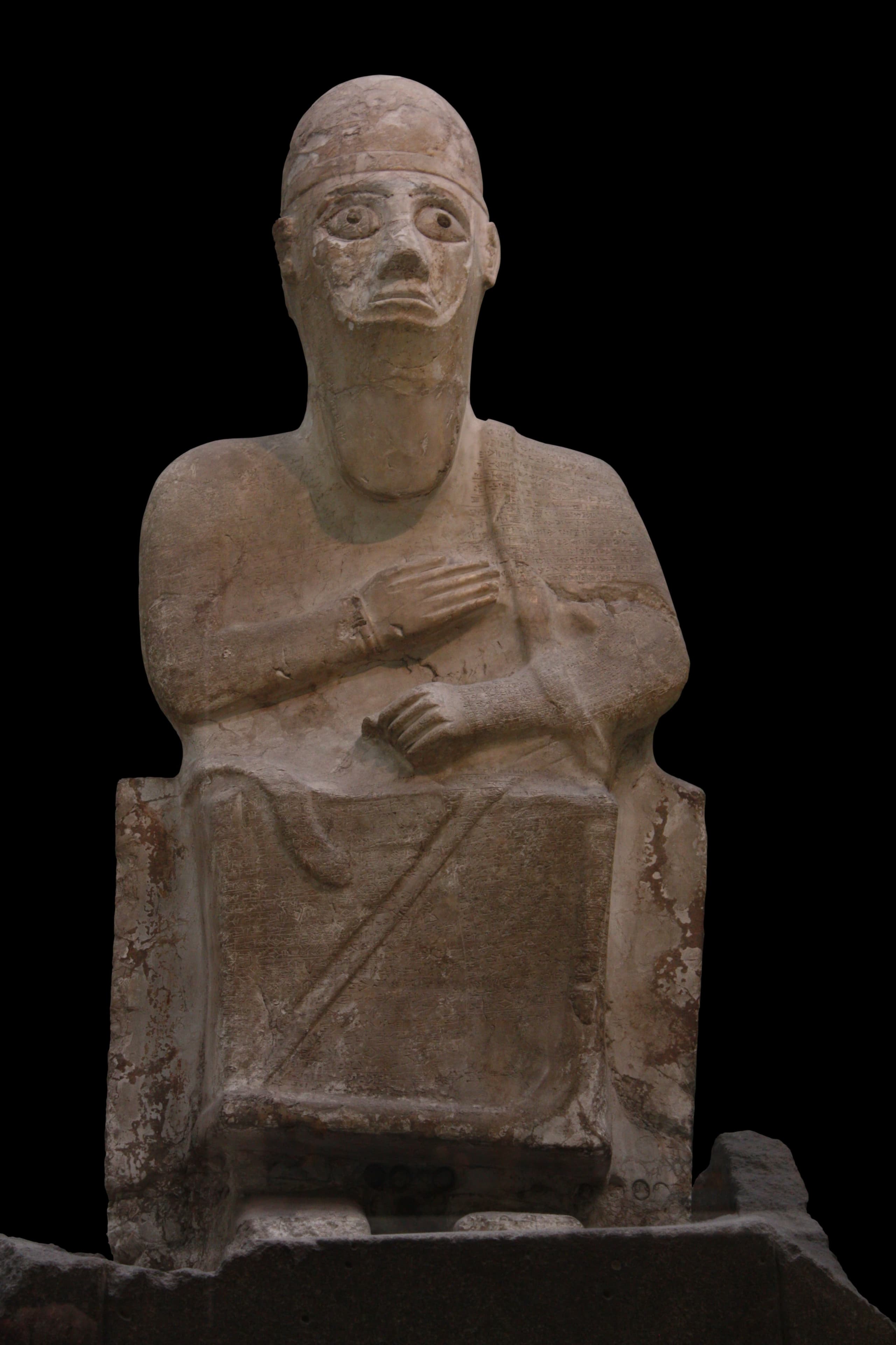 A magnesite statue of a man in royal attire, holding a cup and standing with one hand raised as if giving orders or making declarations. The long inscription is inscribed on the front of his skirt