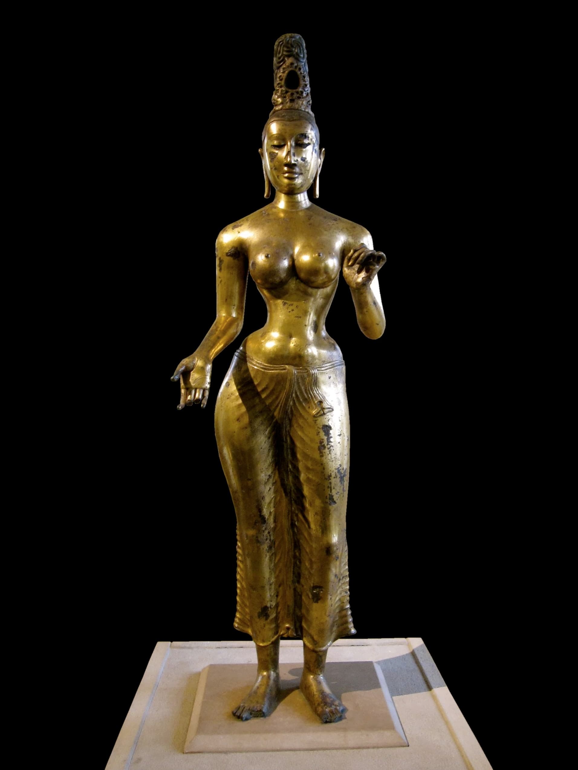 A standing bronze figure of a goddess, nearly life-size, with one hip shifted and traces of gold visible on the surface. The figure wears jewellery and a lower garment; the torso and limbs are bare.
