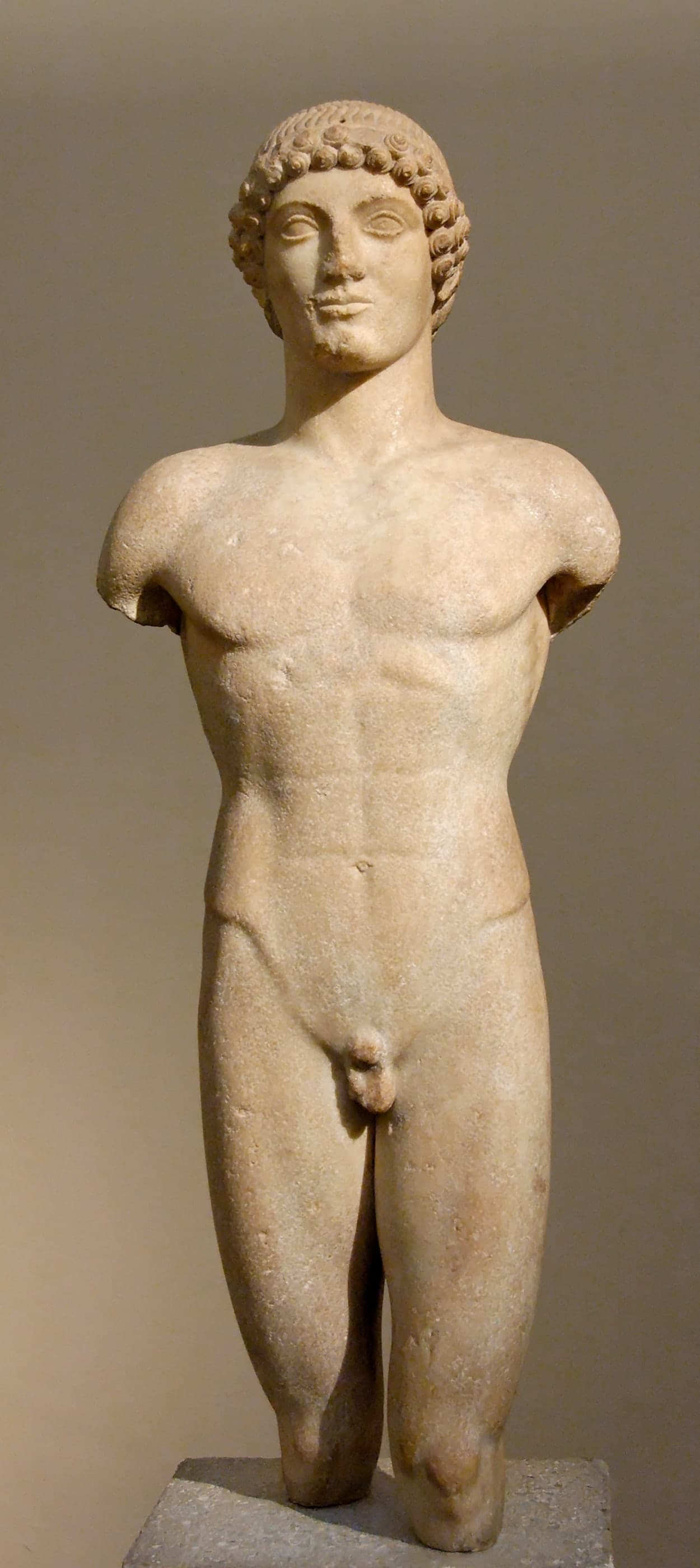Marble statue of a nude standing youth in frontal pose, head and torso preserved, arms and lower legs missing, hair carved in neat Archaic rows.