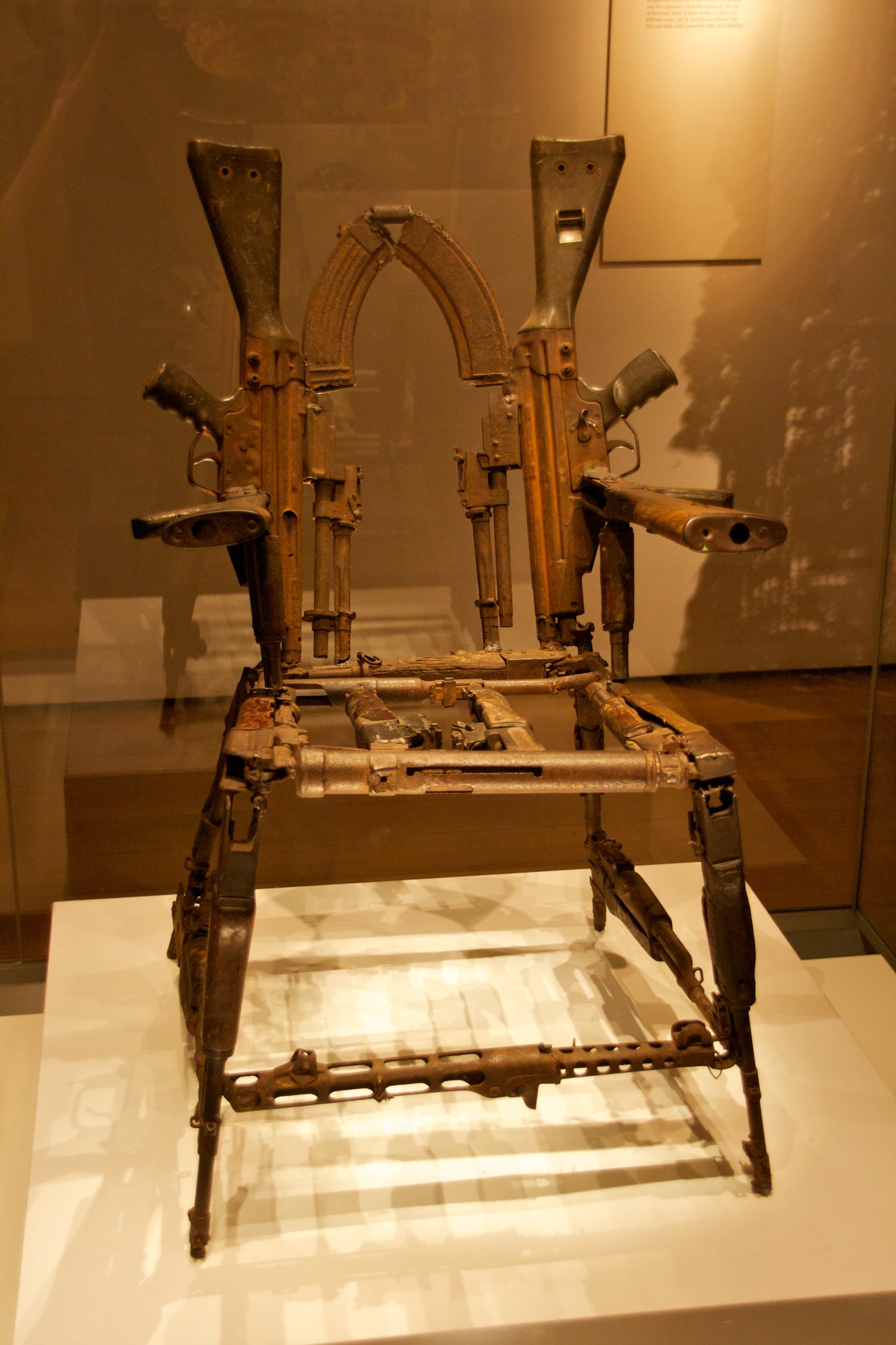 A high-backed chair constructed from welded rifle components: wooden stocks form the seat and back, metal barrels curve into arms and legs, and trigger mechanisms are visible in the joints.