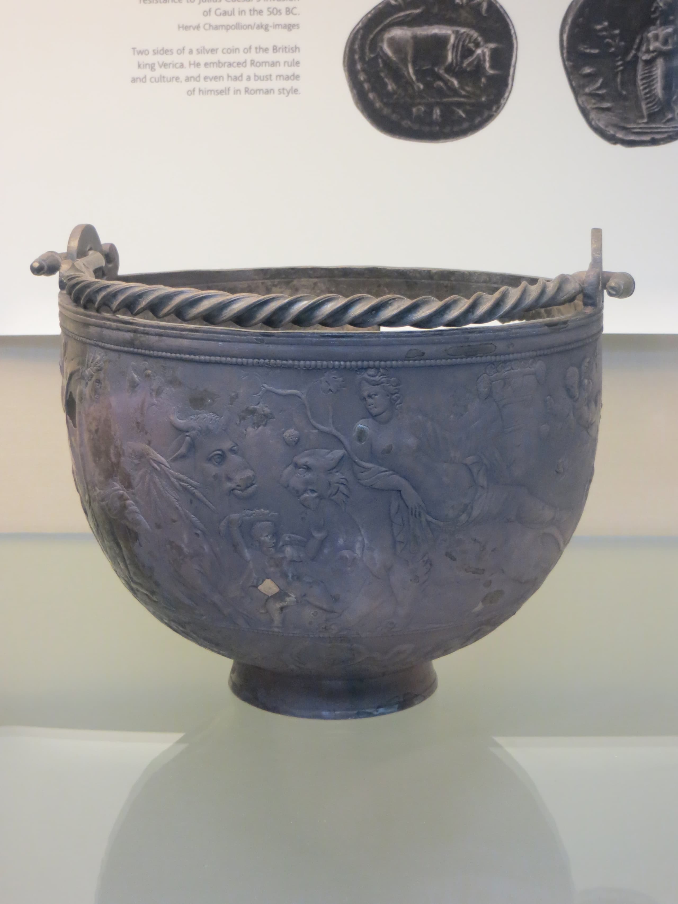 A small hemispherical silver vessel with a curved, twisted handle and engraved figures of women and animals arranged around its upper edge.