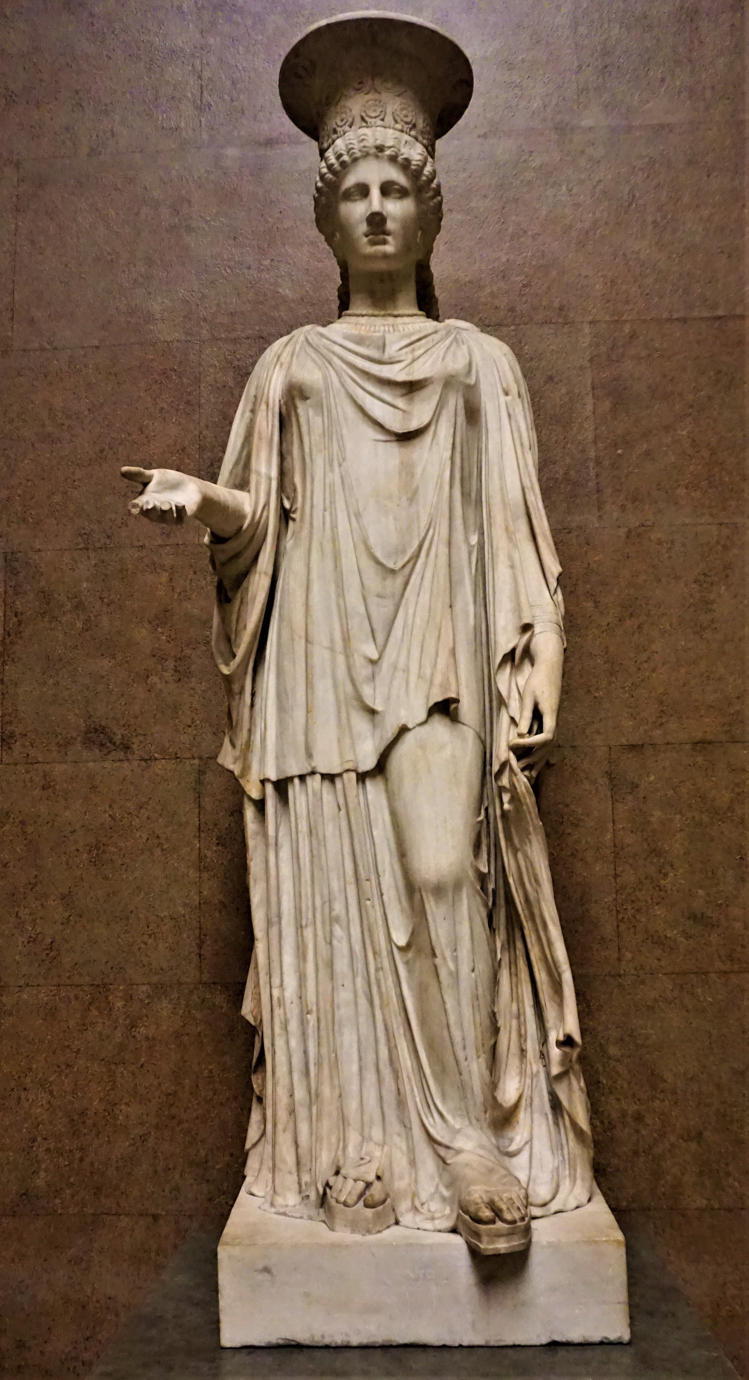 A full-length marble female figure standing frontally in heavy draped garments, a tall cylindrical headdress on her head, carved as a supporting column rather than a free pose.