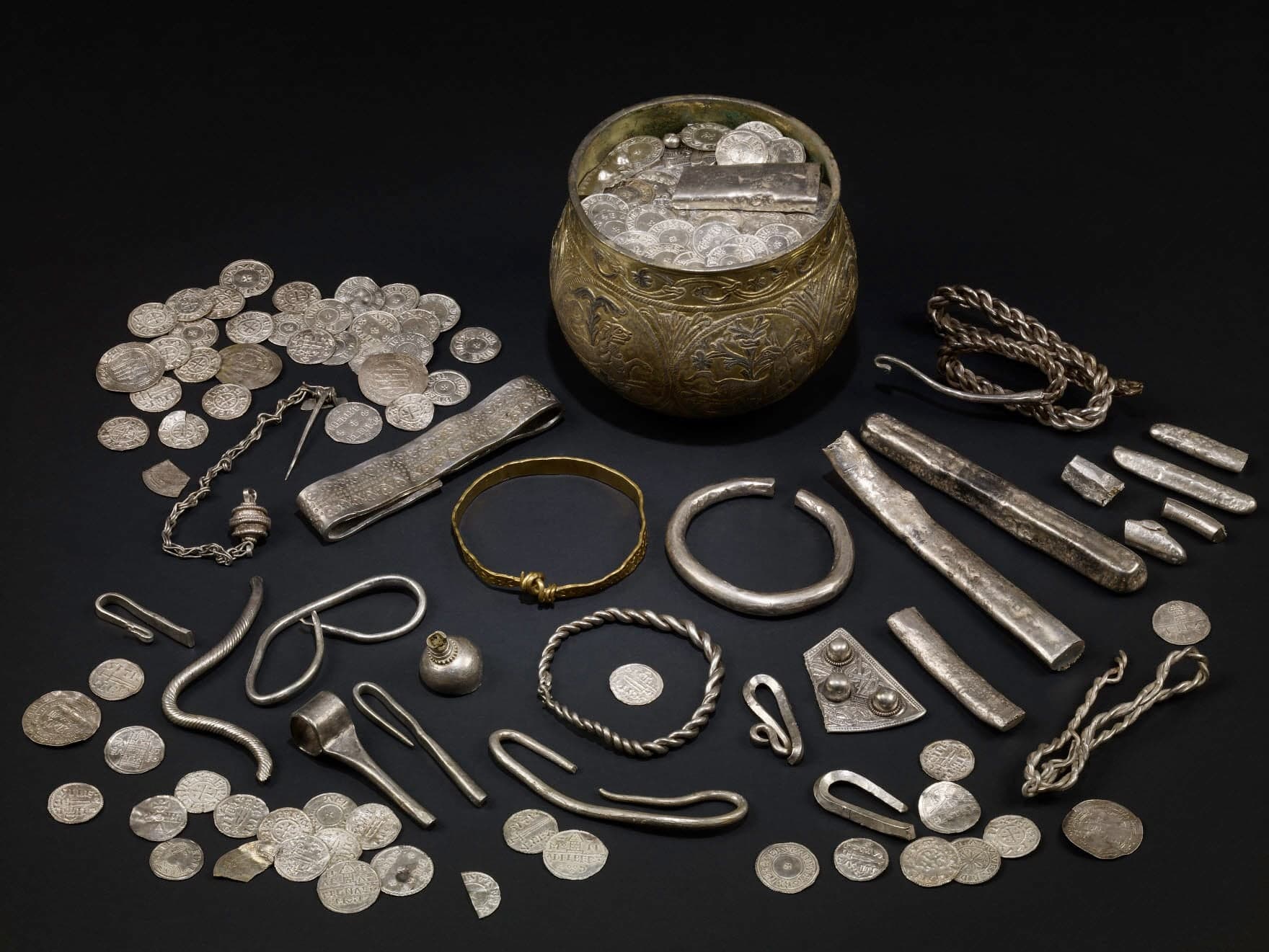 A collection of silver coins, arm-rings, and ingots displayed alongside a decorated gilt silver cup, all items tarnished dark grey with patches of lighter metal showing through.
