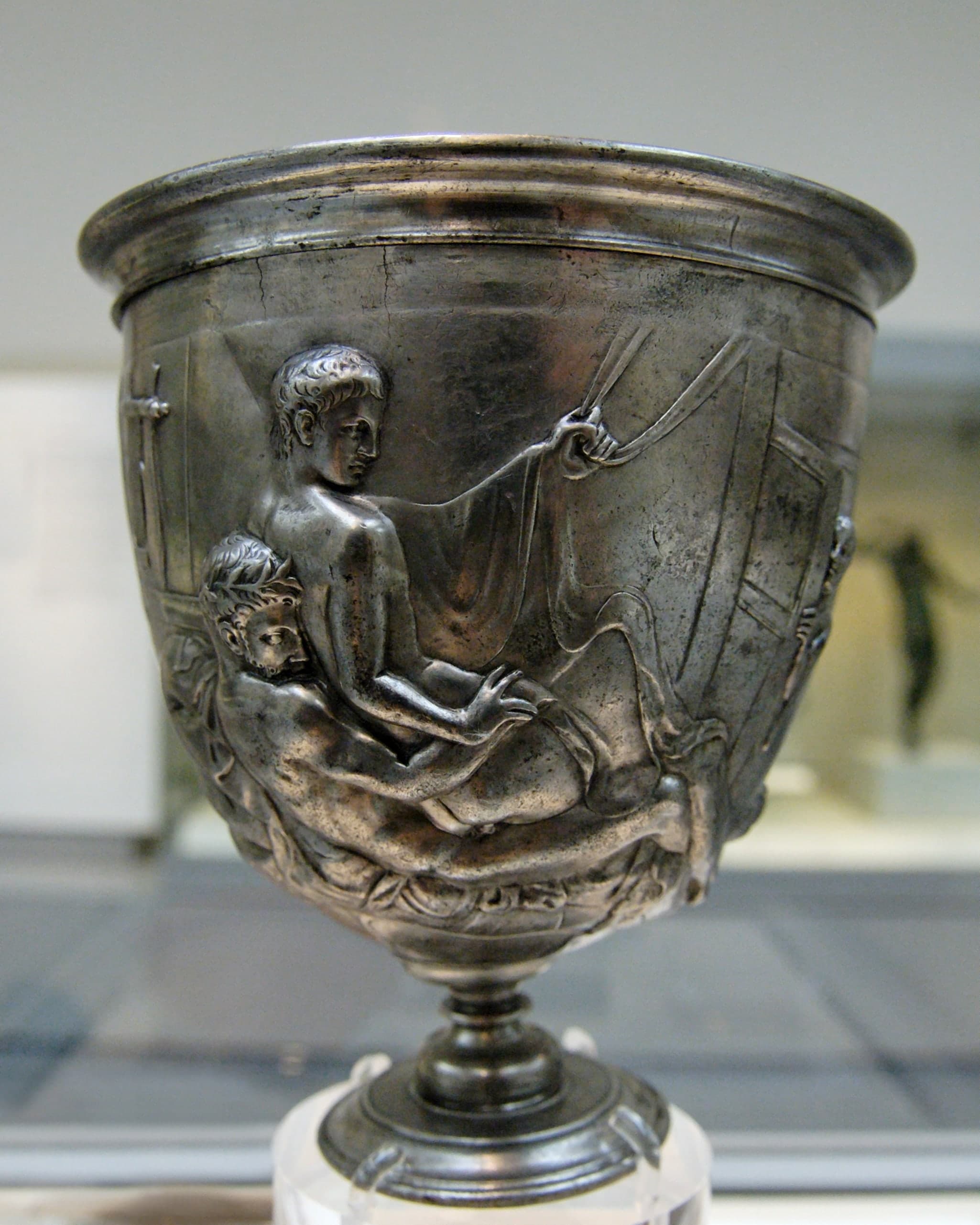 A cylindrical silver drinking cup with two raised relief scenes showing male couples in explicit sexual acts, each scene occupying one side of the vessel.