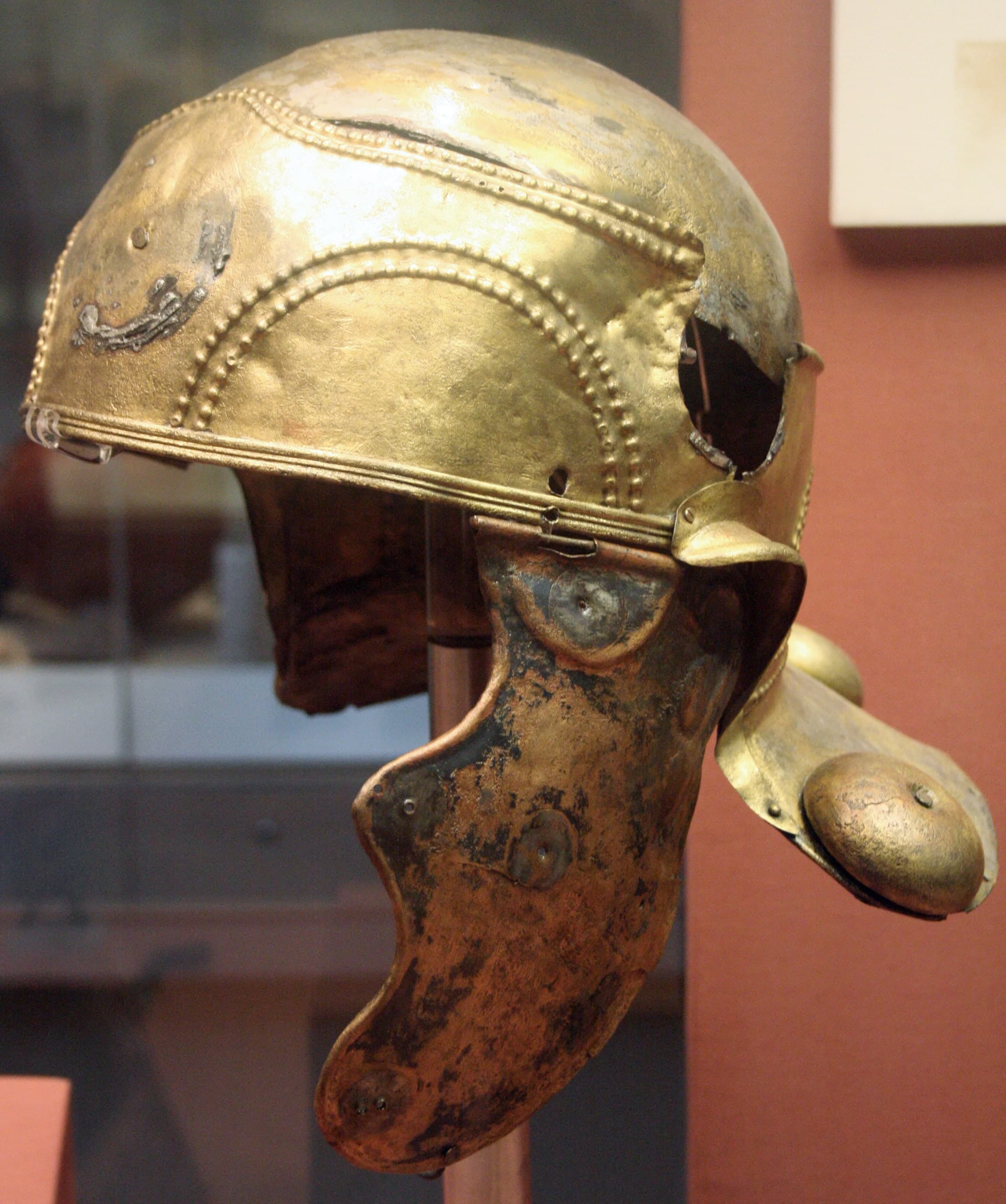 A copper-alloy Roman helmet casing with rounded cap, flaring neck guard and hinged cheek pieces, showing three hollow bosses on the exterior and a worn, mottled green-brown surface.