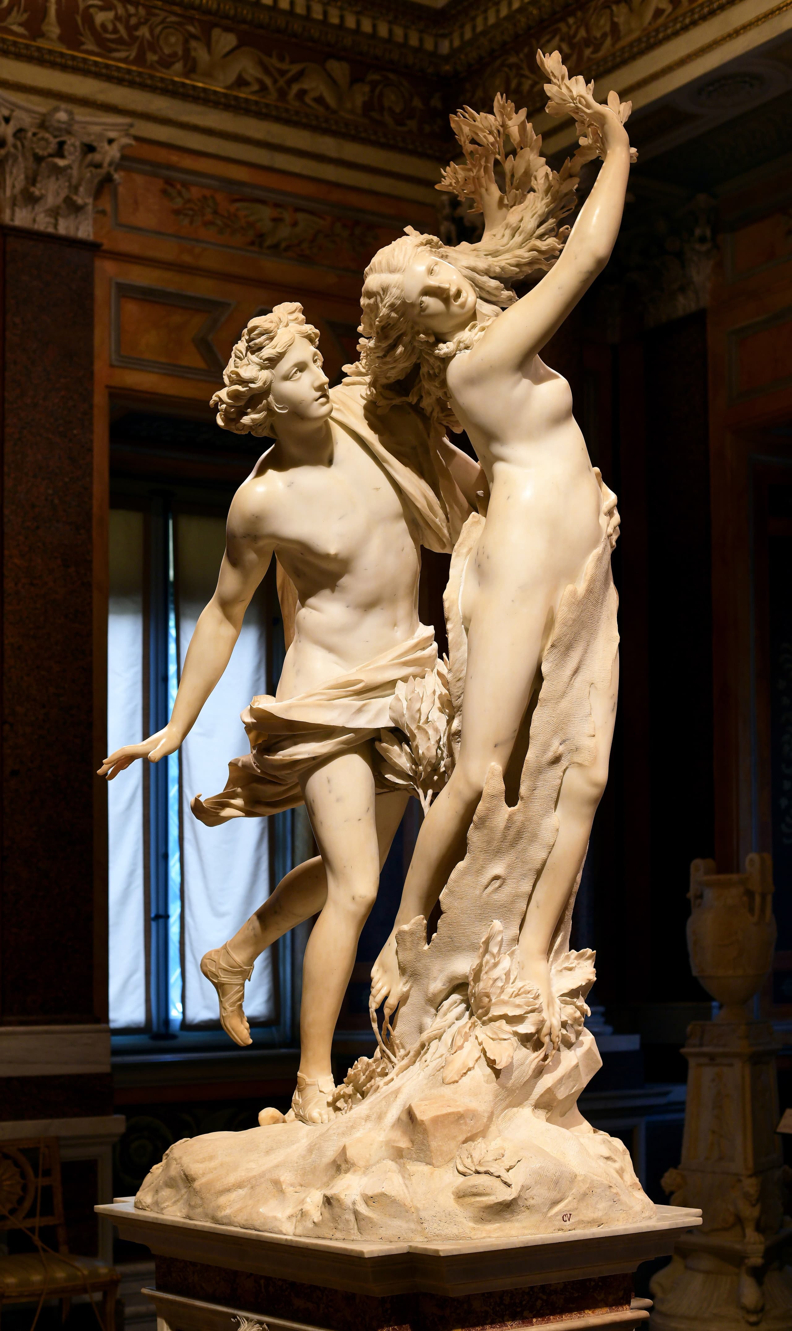 Two pale marble figures in a rising twist: a youth strides and reaches from behind as a young woman turns away, her fingers and hair opening into leaves while bark spreads across her body.
